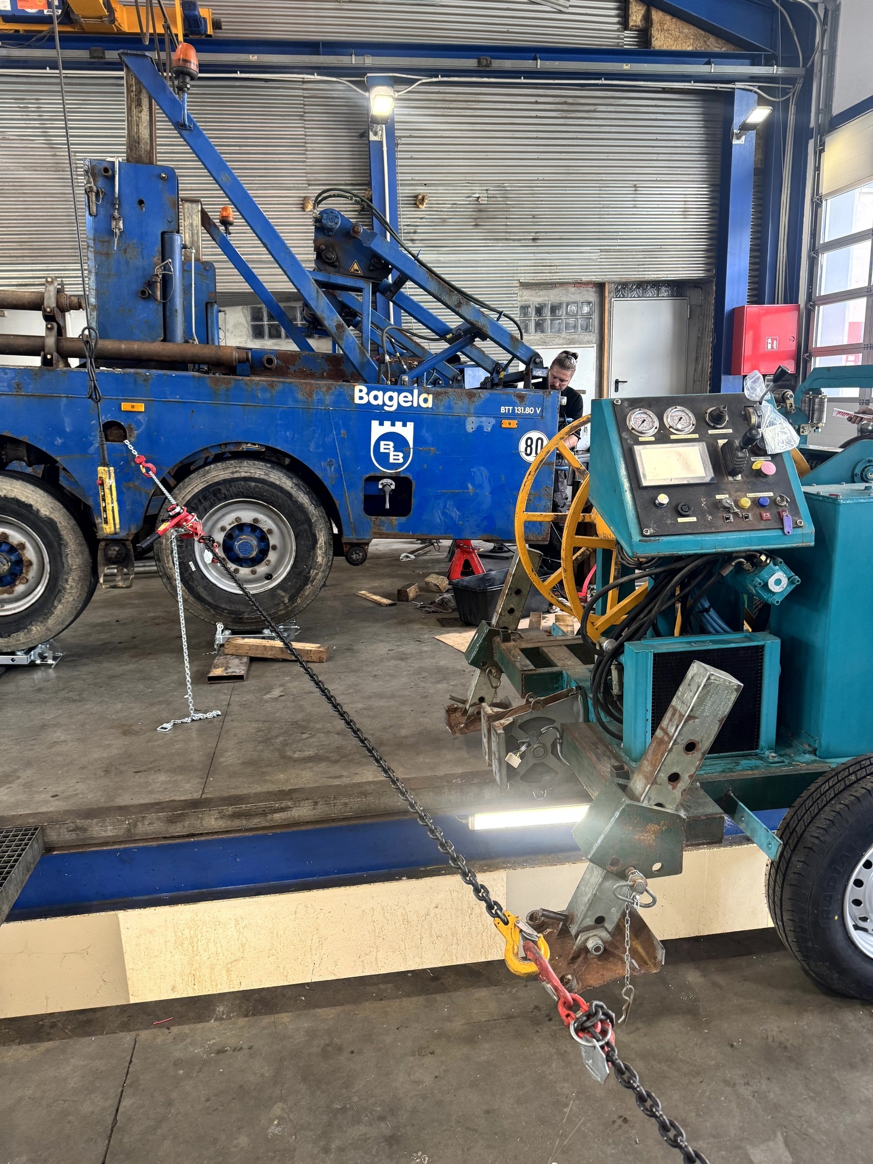 Bagela capstan cable-pulling winch in a workshop—mobile unit with control panel and gauges, chained for load testing to a vehicle/anchor; service, inspection and repair for utility line installation.